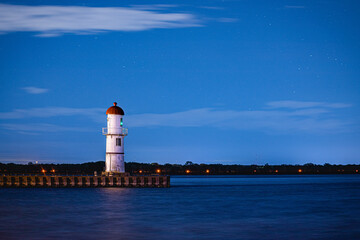 The image shows a lighthouse in the darkness of the night, captured with a long exposure. The clear...