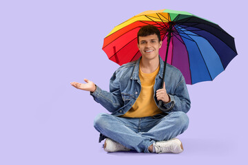 Young man with rainbow umbrella sitting on lilac background