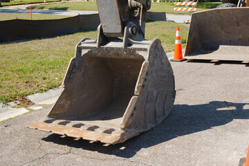 Close-up of an excavator bucket made of steel, with visible teeth along the edge for digging. It is positioned on a paved surface with a construction site or work area visible in the background, indic © Del Harper