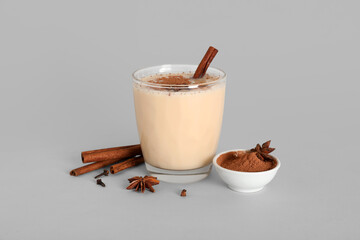 Glass of tasty eggnog cocktail with cinnamon, star anise and bowl of cocoa powder on grey background