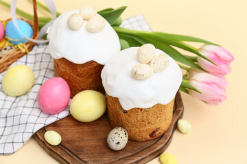 Wooden board with sweet Easter cakes, eggs and beautiful tulip flowers on yellow background