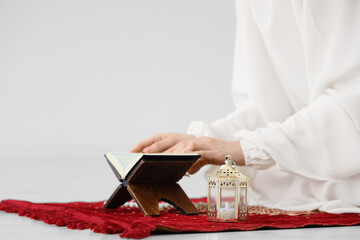 Obraz premium Muslim woman with fanoos and Koran praying on prayer carpet at home, closeup. Ramadan celebration