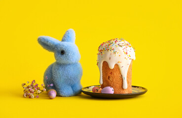 Plate with sweet Easter cake, painted eggs, toy bunny and gypsophila flowers on yellow background