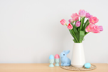 Vase with bouquet of tulip flowers, bunny and painted eggs on table near white wall, closeup. Easter celebration