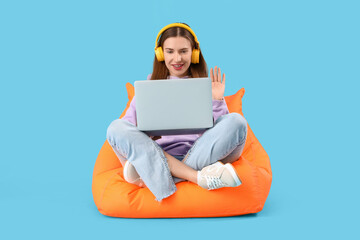 Stylish young woman in hoodie with headphones and laptop video chatting on beanbag against blue...