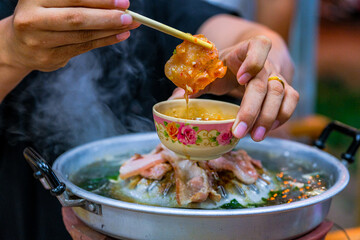 Delicious Close-Up of Asian Cuisine with Hands Holding Chilled Meat Dumpling and Coconut Sauce Over Hot Pot in a Vibrant Setting