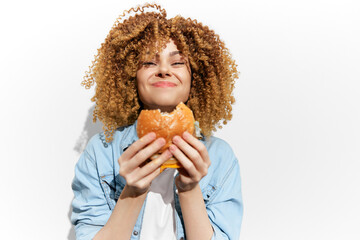 Young woman with curly hair joyfully enjoying a delicious burger against a bright white background,...