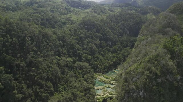 Drone flies down valley with Cahabon River cutting through the Semuc Champey National Park, as camera pans down over iconic pools on sunny afternoon in Guatemala