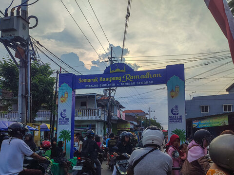 A lively street scene at the entrance of "Semarak Kampung Ramadhan" in Tambak Sedati, Surabaya. The sky is dramatic with clouds, while people on motorcycles and pedestrians fill the bustling road.