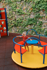 Little girl stands on a colorful carousel holding a handrail near a stone fence entwined with ivy