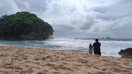 A father and son enjoy a summer walk on the sandy beach along the ocean under a sky dotted with clouds