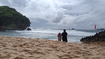 A father and son enjoy a summer walk on the sandy beach along the ocean under a sky dotted with clouds