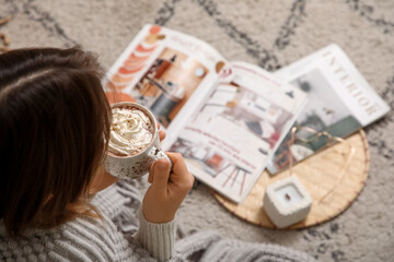 Young woman with cup of hot chocolate and whipped cream reading magazine on floor at home, top view