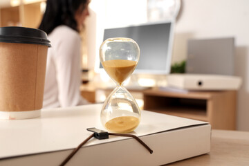 Hourglass with coffee cup on folder against businesswoman working in office, closeup