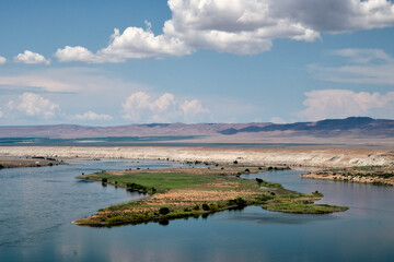 A beautiful view of a lush green island surrounded by a serene river, set against a backdrop of rolling hills and a blue sky dotted with fluffy clouds.