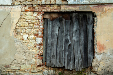 The doorway in the brick house is boarded up. The brick wall of the old house is partially destroyed. The door is blocked with old boards. Green grass at the foot of the brick wall.