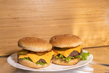 Two cheeseburgers with beef patties, cheddar cheese, pickles, lettuce, and tomatoes on sesame buns, served on a white plate with a rustic background.
