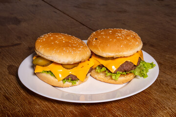 Two cheeseburgers with beef patties, melted cheese, lettuce, and pickles on sesame buns, served on a white plate atop a rustic wooden surface.