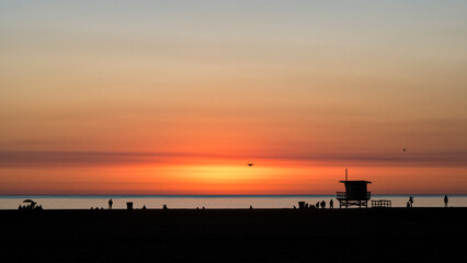 Obraz premium A stunning sunset paints the sky in vibrant hues, casting silhouettes of a lifeguard tower and beachgoers, capturing the essence of relaxation and the beauty of nature.