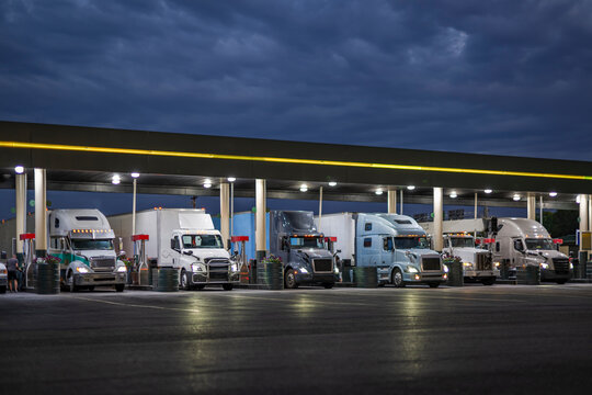 Different big rig semi trucks with turned on headlights and loaded semi trailer standing on the truck stop gas station at night time fill up the trucks tanks to continue their routes in the morning