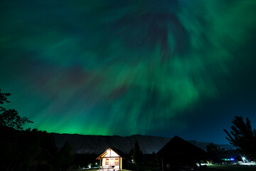 A captivating view of a cozy cabin illuminated by the spectacular display of the aurora borealis,...