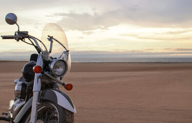 A lone motorcycle stands on the sandy surface of the Namib Desert against a dramatic cloudy sky. The sunset lighting highlights the contrast between nature and machine.