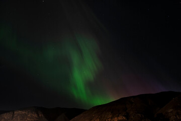 The Northern Lights casting an enchanting glow over the dark mountain silhouette, creating a mesmerizing contrast that draws viewers into a dreamlike, celestial experience.