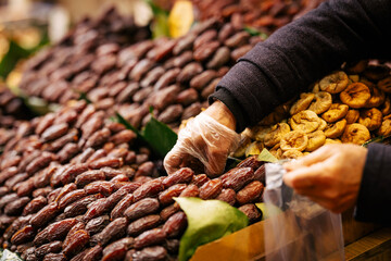 A vendor wearing gloves selects fresh dates at a traditional market. Stacks of dried fruits,...