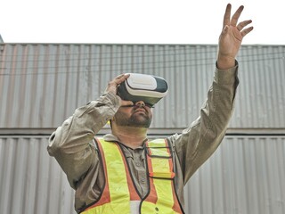 A construction worker wearing safety gear and a virtual reality headset, gesturing while interacting with a virtual environment in an industrial warehouse or manufacturing facility.