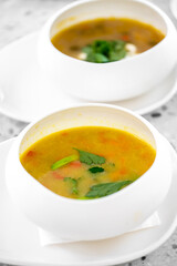 A close-up of two elegant bowls of soup, one in the foreground with vibrant yellow broth and greens, and the other in the background, featuring a rich, darker color.