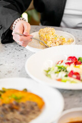 A close-up of a person's hand holding a fork, about to take a bite of a delicious dish. In the foreground, a beautifully presented salad and main course plate are visible.