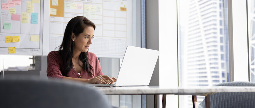Positive focused engaged Latin business leader woman typing on laptop in office workspace alone, working on project strategy, effective management, sitting at computer. Low angle, banner shot