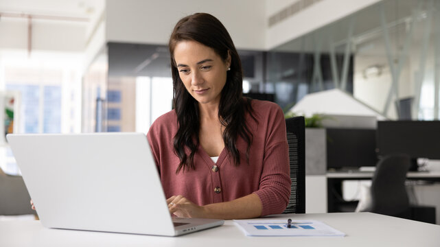 Focused positive business professional woman working on project in office alone, typing on laptop at paper marketing report, developing creative strategy, writing plan for effective management