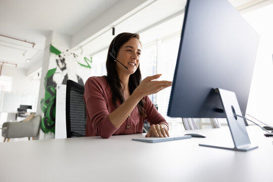 Positive female support center manager in headphones with mic speaking to client at computer. Business coach, leader, blogger giving online training webinar on Internet. Low angle shot