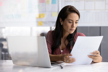 Latin business professional woman reading paper financial report at workplace, checking legal...
