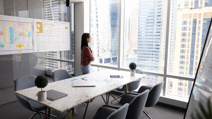 Thoughtful young business leader woman standing alone in office meeting room, looking out of window at urban buildings, city view, thinking on project marketing strategies, company growth
