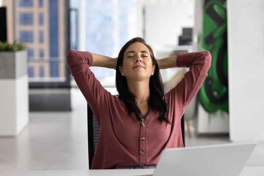 Peaceful positive Hispanic manager woman breathing fresh air at work table, relaxing on comfortable office chair, stretching body, enjoying break, leisure, completed project, smiling with closed eyes