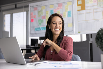 Cheerful Hispanic business project manager woman sitting at large workplace table in office space,...