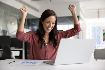 Happy Latin businesswoman making winner hands at laptop, laughing, feeling excited, overjoyed,...