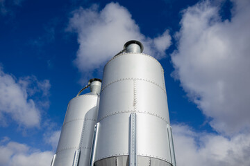 Two large silver industrial silos stand against a bright blue sky with scattered white clouds, reflecting sunlight. The cylindrical storage structures feature rivets, pipes, and ventilation systems © ugis