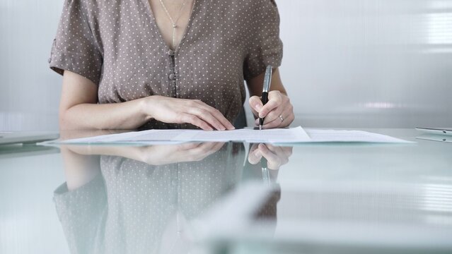 Businesswoman wearing brawn dotted blouse is signing a contract at a sleek glass desk in a modern office, closeup. Business people concept