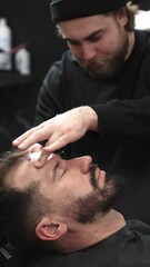 Fototapeta premium Professional barber carefully shaving client's beard with straight razor, applying gel and using hot towel for precise grooming in contemporary barbershop setting, vertical view