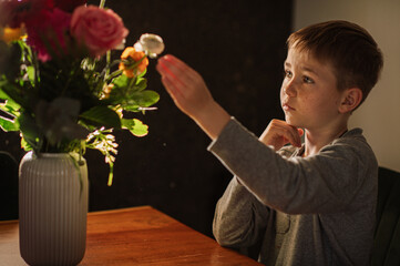 A little boy admires a bouquet of flowers in a vase on the table at home, gently touching the...