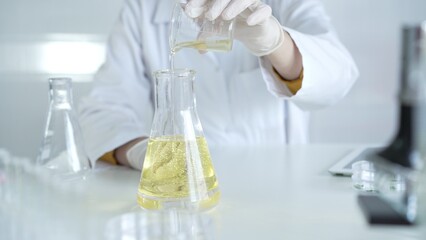A scientist, wearing a lab coat and white protective gloves, is pouring a yellow oily liquid from one beaker to another in laboratory, close up. Medicine and science concept