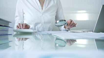 Businesswoman analyzing financial data with magnifying glass and calculator, performing accounting and auditing tasks in office. Audit and taxes theme in business