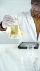 Man scientist wearing white protective gloves, and glasses is holding a yellow chemical solution...