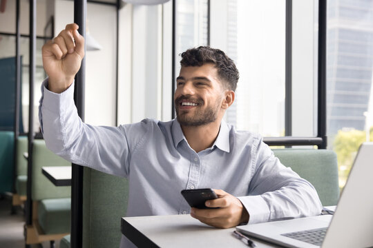 Young man sit at table with laptop in modern cafeteria or coworking area, working alone in public place, raise his arm, waved, calls out to familiar person, colleague for talk or waiter to place order