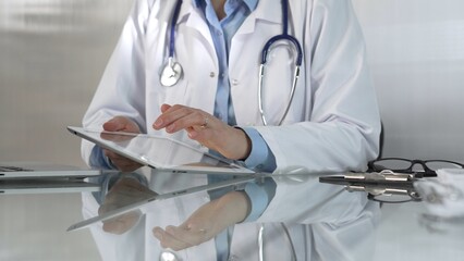 Doctor woman, wearing a blue blouse under a medical coat, is using digital tablet in modern healthcare setting, interacting with patient data, close-up. Medicine concept