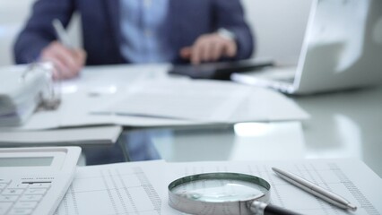 Magnifying glass, calculator, pen, laptop and documents are lying on accountant's desk. Businessman wearing blue suit is working with documents on the background. Business people and audit concept