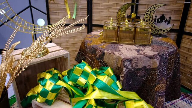 A beautifully decorated Eid display featuring golden Islamic ornaments, ketupat decorations, batik-patterned fabric, and glass jars on an elegant festive table setting.